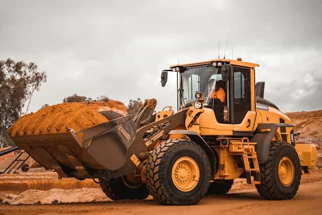 A yellow bulldozer pauses in the midst of reddish sand, driven by someone in an orange uniform.